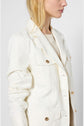 A woman with long blonde hair wears a cream-colored jacket with gold buttons over a beige top, standing against a plain light background. Her face is partially out of frame.