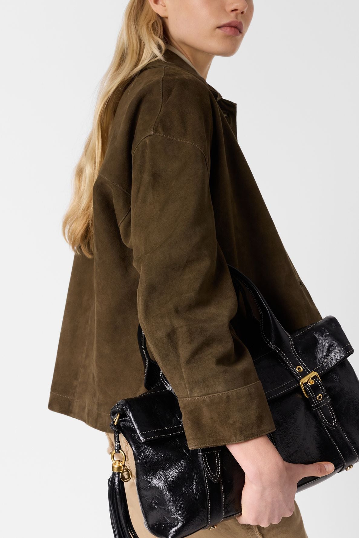A woman with long blonde hair wears a brown veste courte en cuir velours and carries a large black leather handbag with gold hardware over her shoulder, against a plain light background.