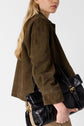 A woman with long blonde hair wears a brown veste courte en cuir velours and carries a large black leather handbag with gold hardware over her shoulder, against a plain light background.