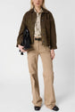A person wearing a brown cuir velours surchemise, a beige shirt, tan pants with frayed sides, and black shoes holds a black handbag against a plain background.