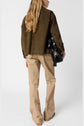Person with long blond hair wearing a veste courte in brown and beige frayed pants, holding a large black handbag, standing with their back to the camera against a plain white background.