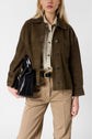 A woman wears a brown veste courte en cuir velours over a beige shirt and tan pants with frayed seams, holding a black leather bag under her arm.