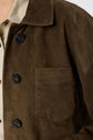 Close-up of a person wearing a brown cuir velours suede jacket with black buttons and a chest pocket, layered over a beige collared shirt. This veste courte is styled so that only the upper torso is visible.