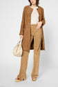 A woman wearing a tan veste sans col suede coat, beige top, patterned tan pants, and platform heels holds a cream handbag. She is standing against a plain light background.