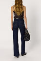 A person with long wavy hair, wearing a satin dentelle black sleeveless top and dark blue jeans, stands facing away while holding a black handbag.