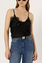 Woman wearing a chic black lace-trim camisole, reminiscent of lingerie chic, paired with high-waisted blue jeans, a black belt with a gold buckle, and round black sunglasses, standing against a plain white background.