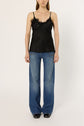 A woman stands against a plain background wearing a black top noir satin camisole with delicate dentelle trim, blue wide-leg jeans, and black shoes. Her arms are relaxed by her sides, effortlessly channeling lingerie chic style.