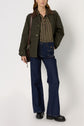A person wearing a green jacket, a chemise à rayures, dark flared jeans, black boots, and carrying a brown shoulder bag stands against a plain background.