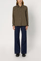 A person stands facing forward, wearing a chemise à rayures—a green long-sleeve button-up shirt with vertical beige stripes—paired with dark blue wide-leg jeans and black shoes. The background is plain and light-colored.