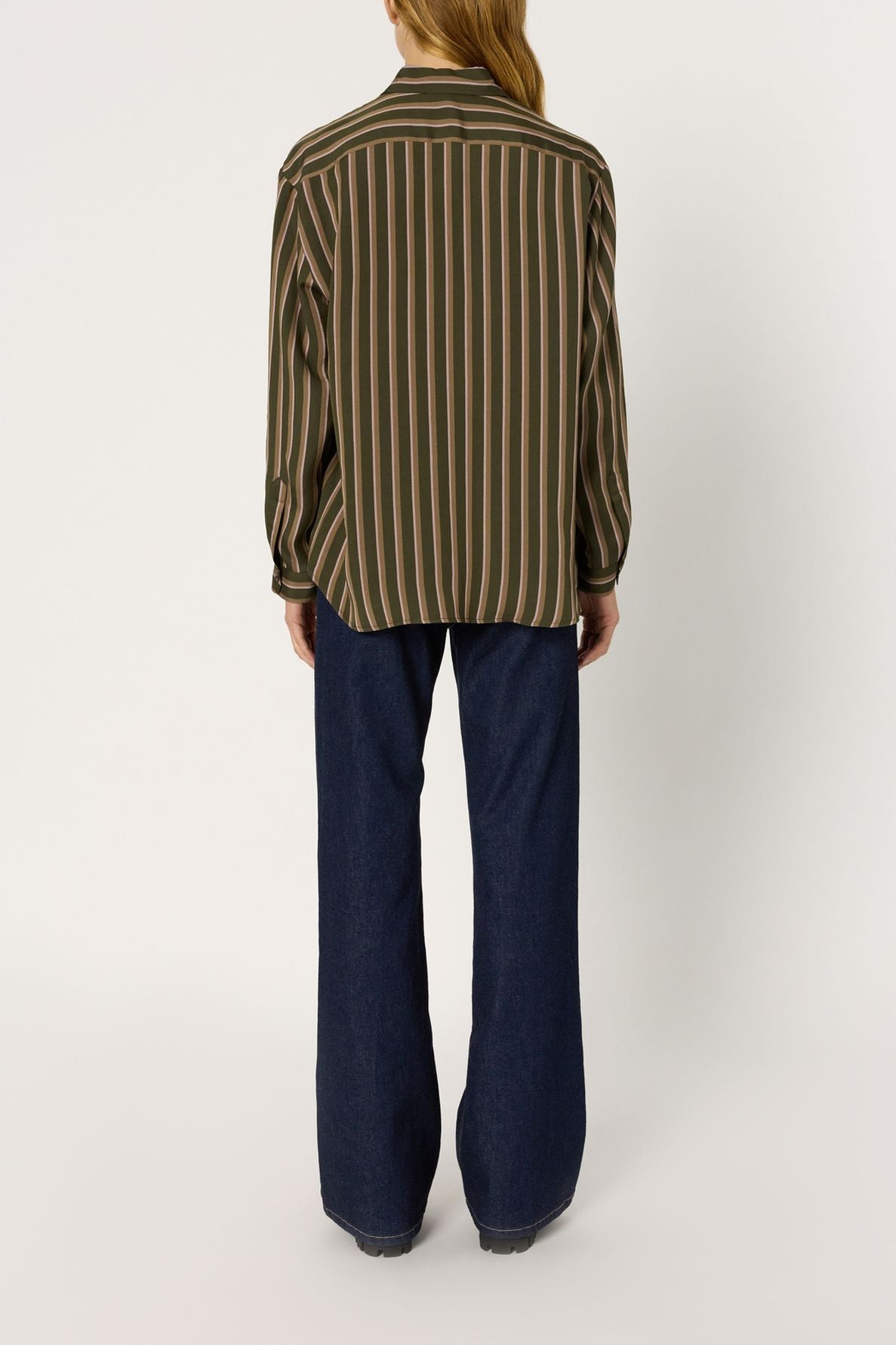 Person standing facing away, wearing a long-sleeve chemise à rayures, featuring green and brown vertical stripes, paired with wide-leg dark blue jeans. Their hair is tied back in a ponytail, and they are standing against a plain white background.