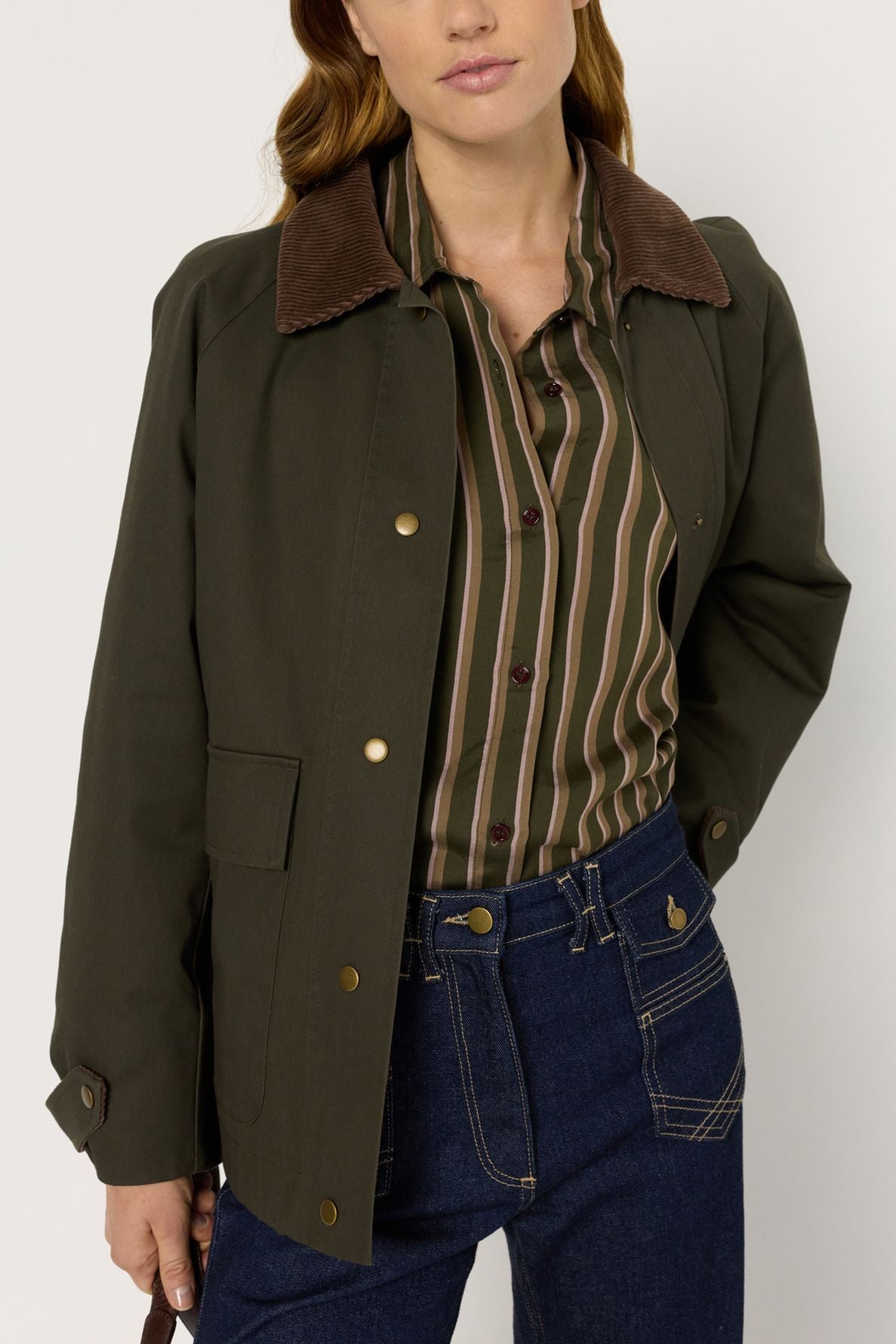 A woman wearing a dark green jacket with a brown corduroy collar, a chemise à rayures button-down shirt, and dark blue jeans stands against a plain light background.
