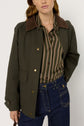 A woman wearing a dark green jacket with a brown corduroy collar, a chemise à rayures button-down shirt, and dark blue jeans stands against a plain light background.