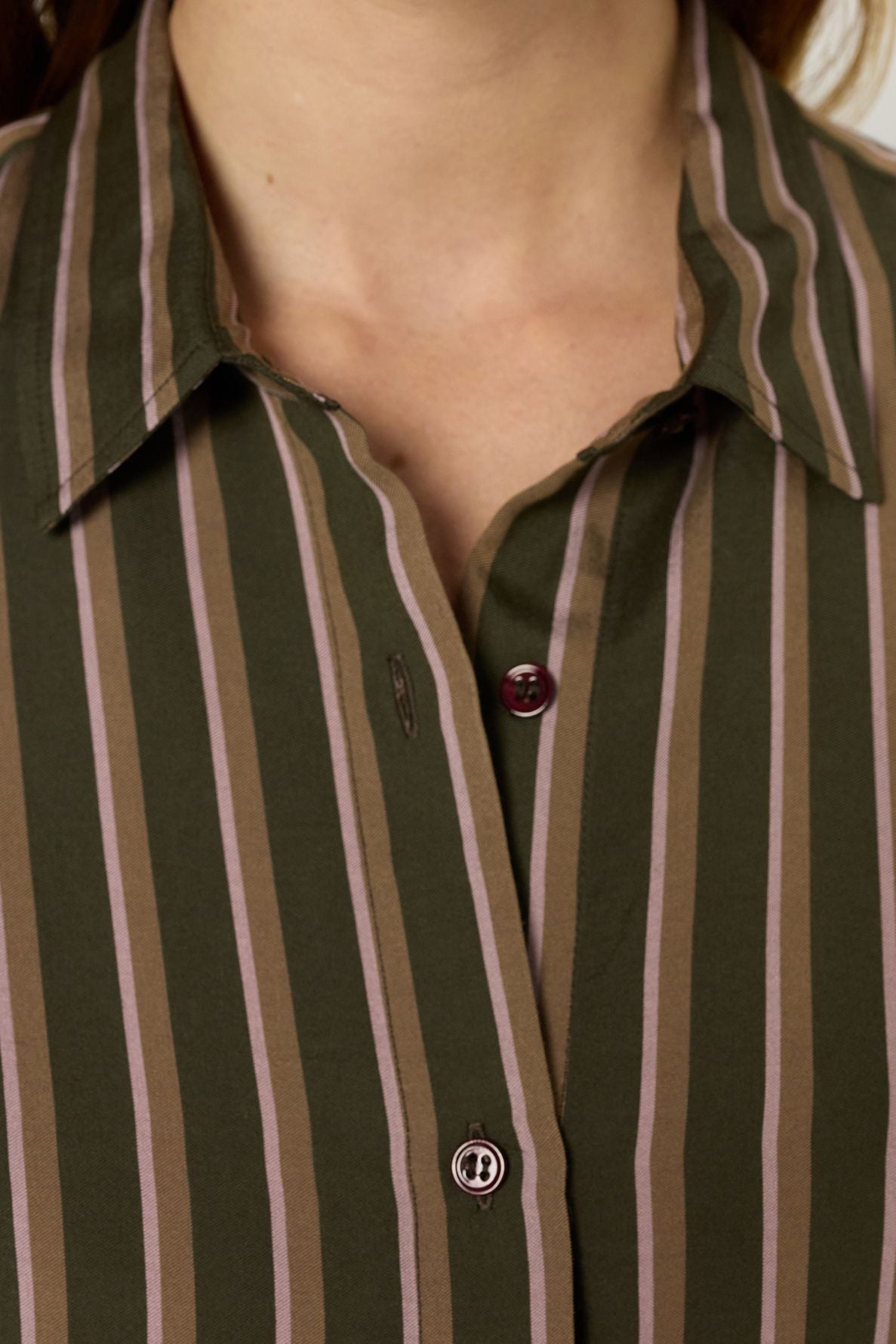 Close-up of a person wearing a chemise à rayures, featuring vertical pink and brown stripes on a green shirt. The chemise kaki has a collar and two visible buttons, with the top button left undone. The image is cropped at the neck and chest.