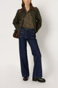 A person stands wearing a green jacket with a brown collar, a chemise à rayures, wide-leg blue jeans, and black shoes, holding a dark brown bag over one shoulder.
