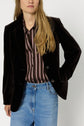 A person with long blonde hair wears a black blazer over a chemise à rayures and blue jeans, standing against a plain white background.