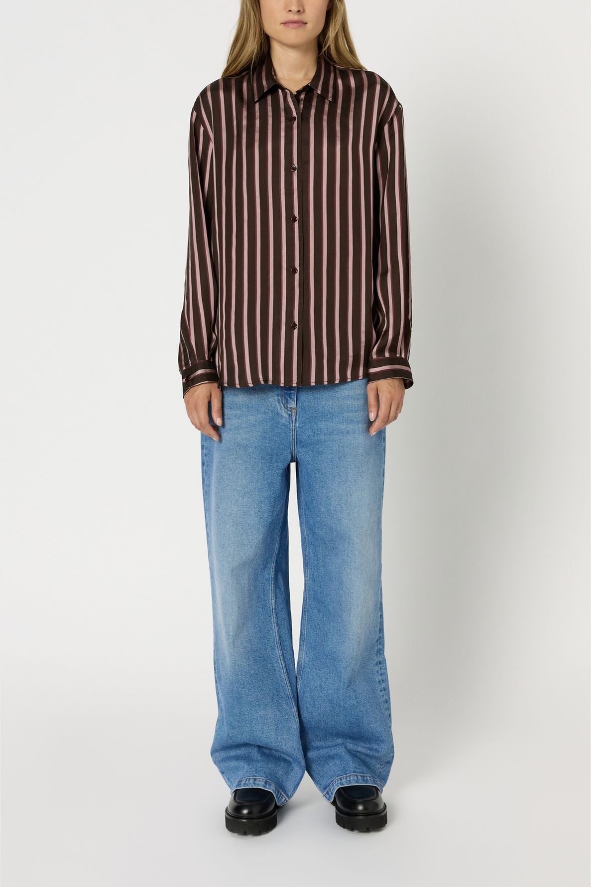 A person wearing a brown and pink vertical striped chemise à rayures button-up shirt, loose blue jeans, and black shoes stands against a plain white background.