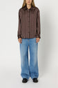 A person wearing a brown and pink vertical striped chemise à rayures button-up shirt, loose blue jeans, and black shoes stands against a plain white background.
