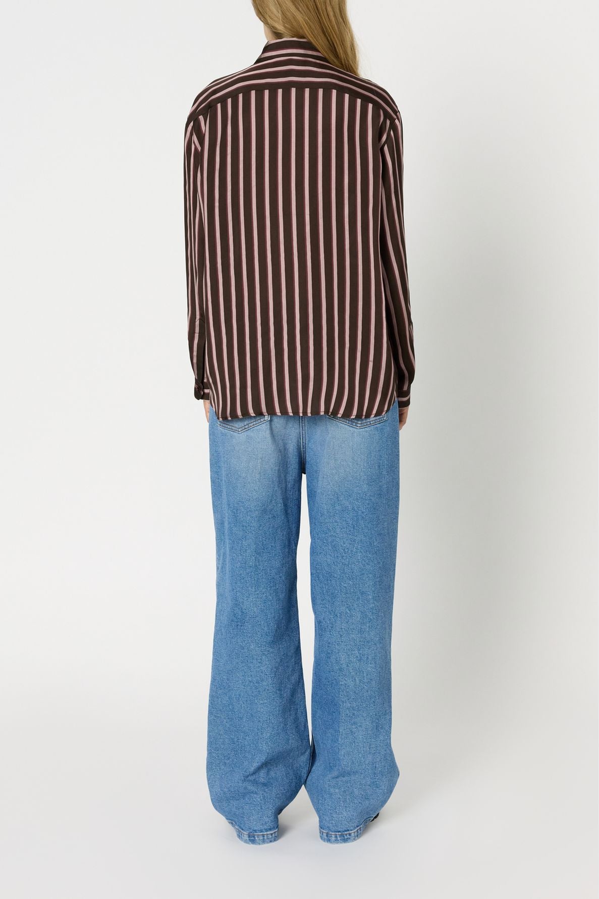 A person stands facing away, wearing a chemise à rayures in brown and pink and loose-fitting light blue jeans, against a plain white background.