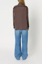 A person stands facing away, wearing a chemise à rayures in brown and pink and loose-fitting light blue jeans, against a plain white background.