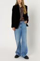 A person wearing a black blazer, a chemise à rayures, light blue wide-leg jeans, and black shoes stands against a plain white background.