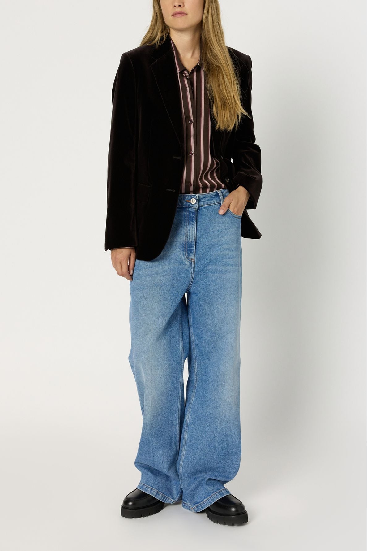 A person wearing a dark velvet blazer, a chemise à rayures button-up shirt, wide-leg blue jeans, and black shoes stands against a plain white background.