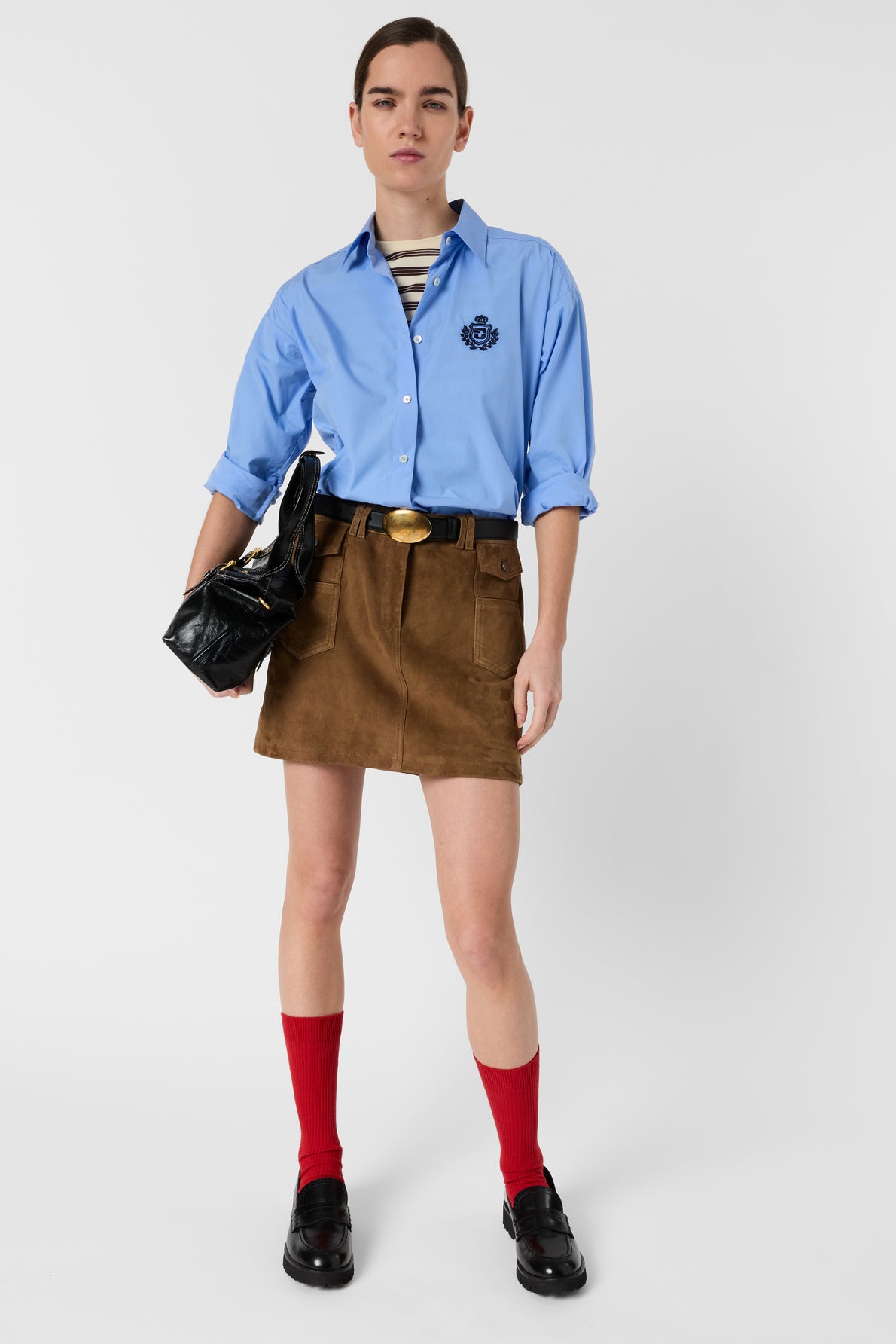 A person wearing a blue button-up shirt, brown suede mini skirt, black belt, black shoes, red socks, and holding a black clutch bag stands against a plain background.