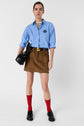 A person wearing a blue button-up shirt, brown suede mini skirt, black belt, black shoes, red socks, and holding a black clutch bag stands against a plain background.