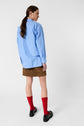 Person with long brown hair wearing a light blue shirt, brown skirt, red knee-high socks, and black shoes, standing and looking over their shoulder against a plain white background.