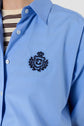 A person wearing a light blue button-up shirt with a dark embroidered crest and F emblem on the chest, over a cream shirt with black horizontal stripes.