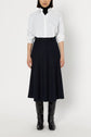 A person stands against a white background wearing a white button-up shirt, black turtleneck, jupe évasée noire mi-longue, and black knee-high boots.