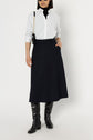 A person wearing a white button-up shirt, black turtleneck, navy jupe midi évasée, knee-high black boots, and a black crossbody bag poses against a plain white background.