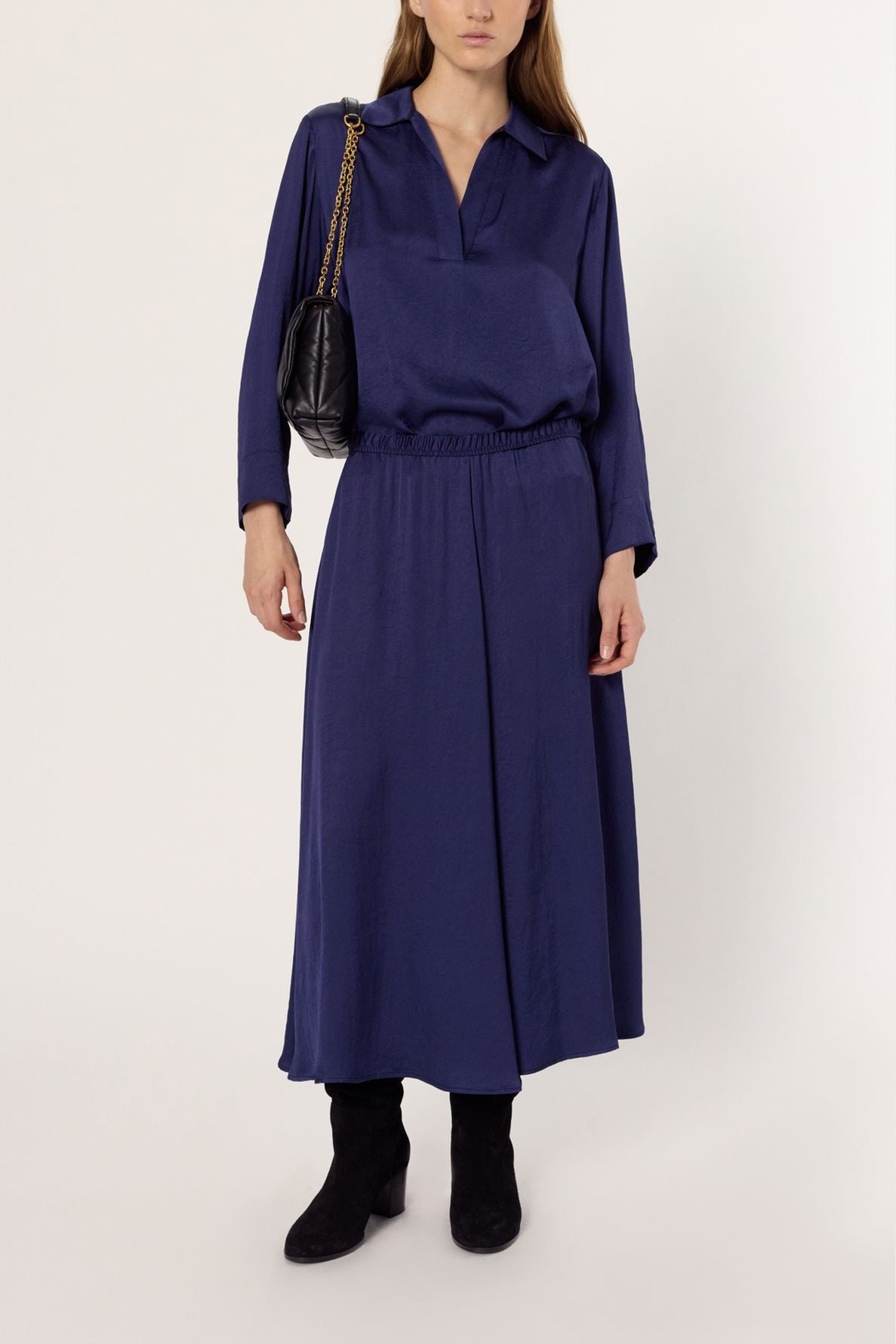 A woman wearing a loose-fitting, long-sleeve navy blue blouse and a matching jupe mi-longue marine, paired with black boots and a black shoulder bag with a gold chain strap, stands against a plain background.