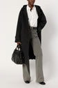 A person wearing a long dark manteau oversize, white button-up shirt, gray flared jeans, and black shoes holds a black handbag.