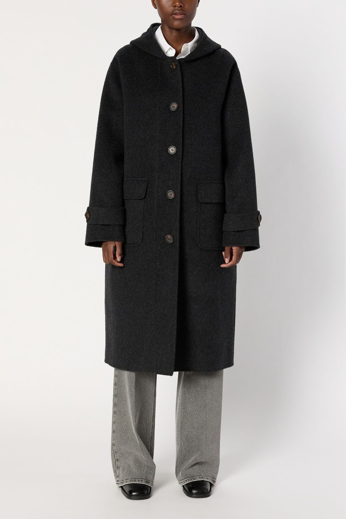 A person wearing a long, dark gray manteau oversize with large buttons, front pockets, and wide sleeves, over gray pants and black shoes, standing against a plain white background.