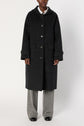A person wearing a long, dark gray manteau oversize with large buttons, front pockets, and wide sleeves, over gray pants and black shoes, standing against a plain white background.