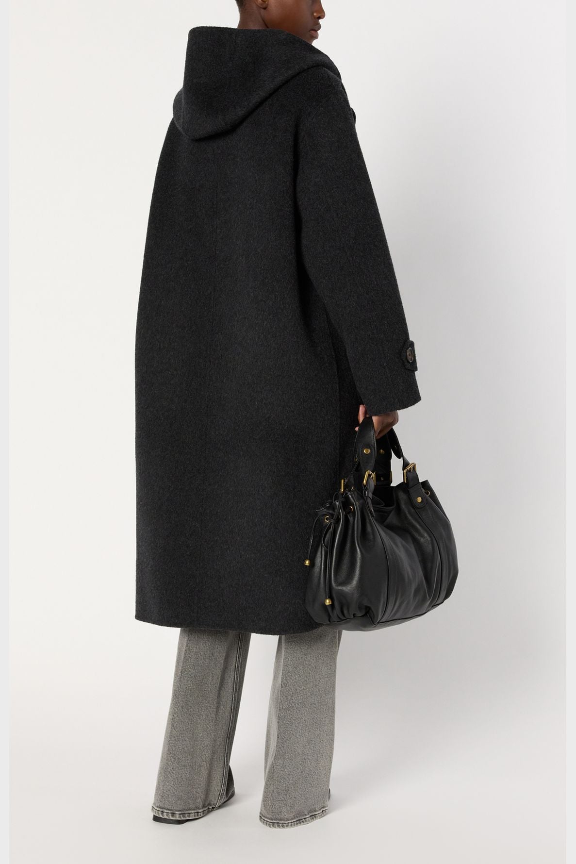 A person wearing a long, dark manteau oversize with a hood and gray pants holds a large black leather handbag. The individual is shown from the back against a plain white background.