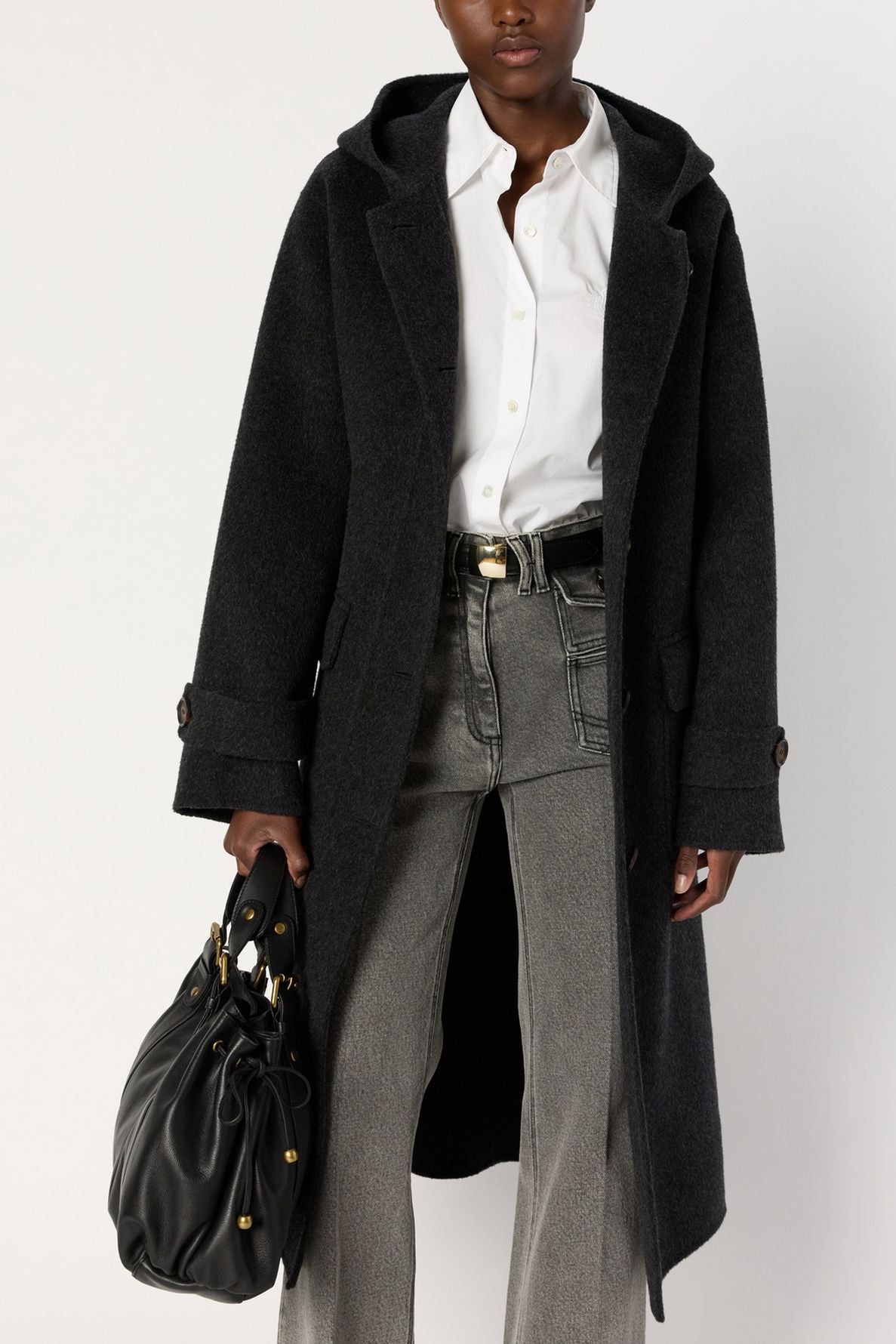A person wearing a long black manteau capuche over a white shirt and gray jeans, holding a black handbag with gold accents.