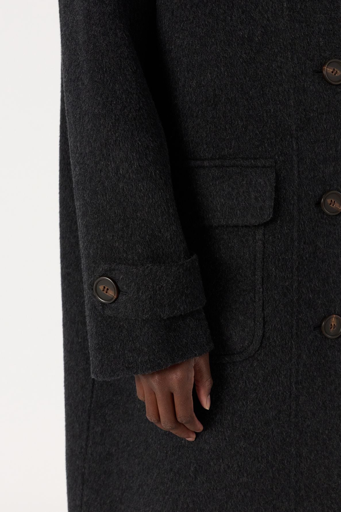 A close-up of a person’s hand resting against the side of a dark manteau laine with large brown buttons and a front pocket. The sleeve also features a buttoned strap detail near the cuff, highlighting the cozy elegance of this timeless coat.