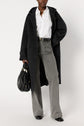 Person wearing a dark manteau oversize, white shirt, gray wide-leg jeans, and black boots, holding a black handbag, standing against a plain white background.