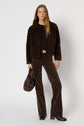 A woman stands against a white background wearing a brown manteau bouclé, a dark brown sweater, patterned brown pants, black boots, and holding a dark brown bag. She has long, wavy hair and a relaxed expression.