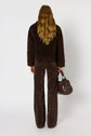 Person with long wavy hair, wearing a brown manteau bouclé textured jacket and patterned brown pants, stands facing away, holding a brown handbag in their left hand against a plain white background.