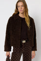 Woman wearing a dark brown manteau bouclé jacket over a dark sweater and brown patterned pants, standing against a plain white background while holding a small clutch bag at her side.
