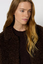 A woman with long, wavy blonde hair wearing a textured dark brown manteau bouclé and a matching sweater, looking slightly to the side against a plain background.