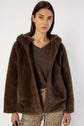 A woman with long dark hair wears a brown manteau court en fausse fourrure over a brown top and leopard print pants, standing against a plain light background.