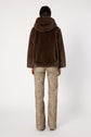 A person stands facing away, wearing a manteau à capuche in brown fausse fourrure and beige leopard print pants against a plain white background.