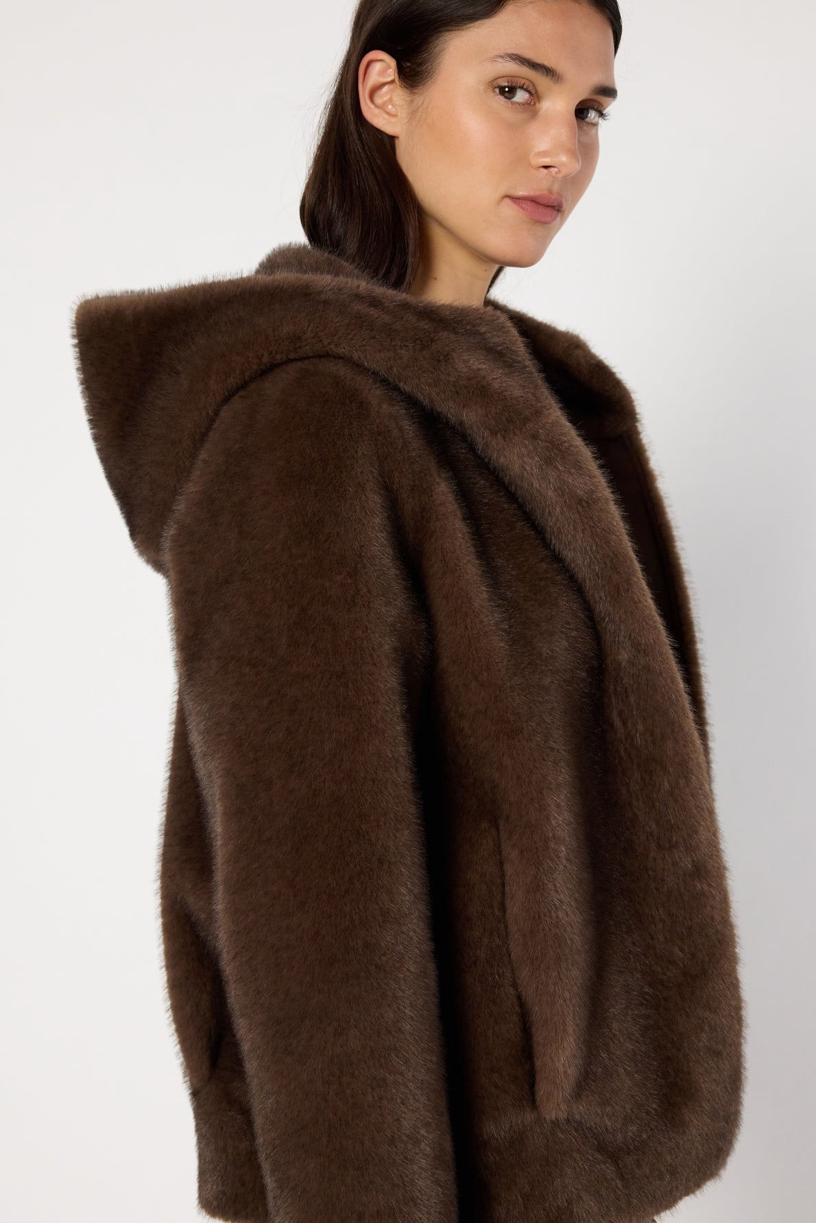 A woman with long dark hair wears a plush brown manteau fausse fourrure à capuche, looking over her shoulder against a plain white background.