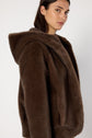 A woman with long dark hair wears a plush brown manteau fausse fourrure à capuche, looking over her shoulder against a plain white background.
