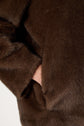 A person’s hand is partially tucked into the pocket of a dark brown, furry manteau fausse fourrure, with only the wrist and part of the hand visible.