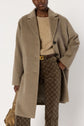 Person wearing a manteau mi-long taupe over a beige sweater and patterned brown pants, accessorized with a brown belt and holding a gold-brown handbag against a plain background.