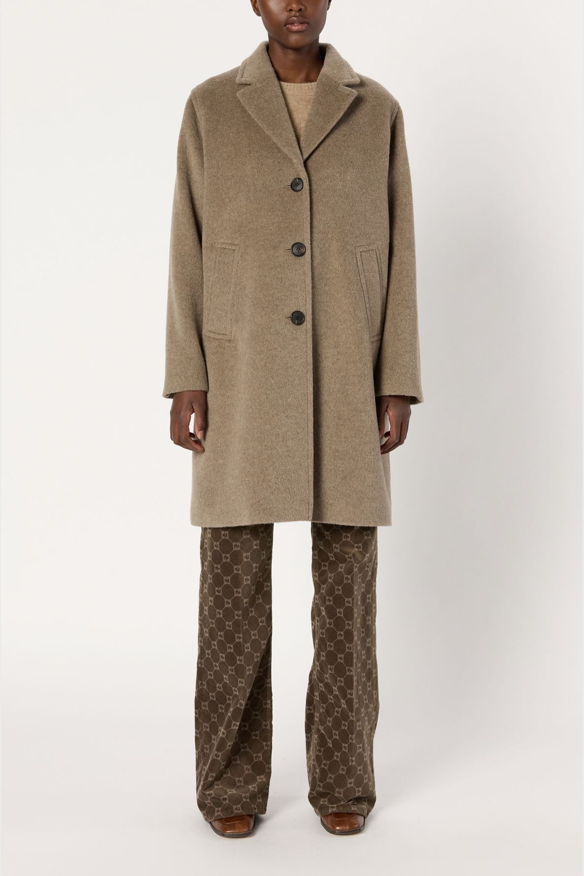 A person wearing a beige manteau mi-long with three buttons, patterned brown pants, and brown shoes stands against a plain white background.